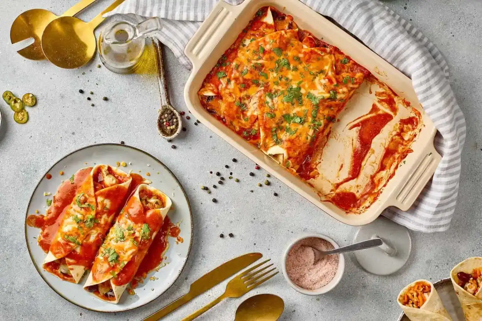 A baking dish of cheesy enchiladas with tomato sauce sits beside a plate holding two enchiladas; gold utensils, seasonings, and napkin are arranged around them.