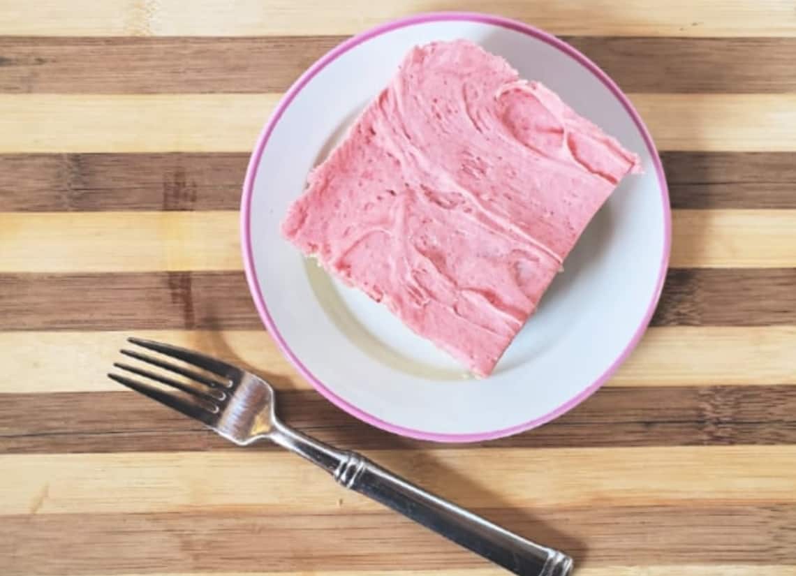 A square piece of cake with pink frosting on a white plate with a pink rim, placed next to a fork on a wooden, striped surface.