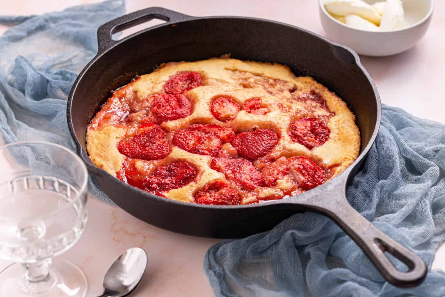A strawberry cobbler baked in a black cast iron skillet sits on a blue cloth, with a spoon, glass, and a bowl of cream nearby.