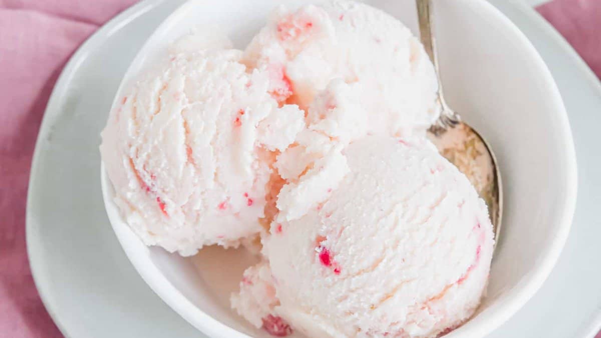A white bowl with three scoops of light pink ice cream, showing small red fruit pieces, and a spoon resting inside.