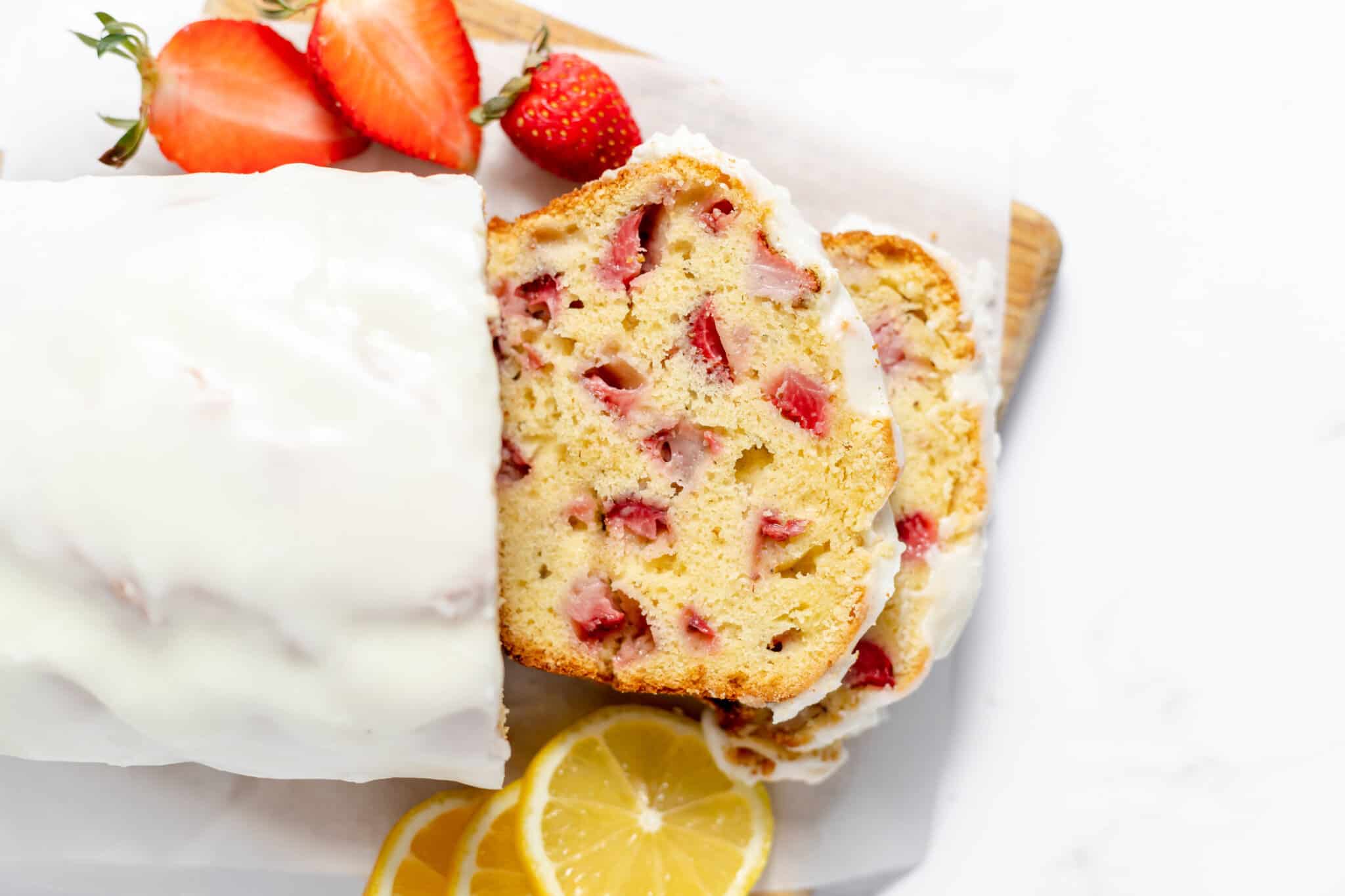 Sliced loaf of strawberry lemon bread with icing, garnished with fresh strawberries and lemon slices on a wooden board.