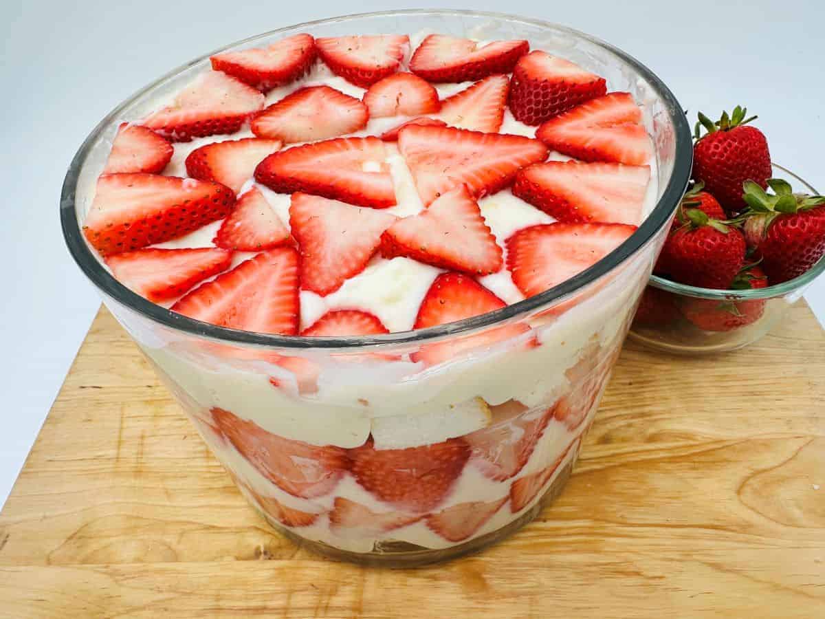 A glass bowl filled with a layered strawberry trifle, topped with sliced strawberries; a small bowl of whole strawberries sits nearby on a wooden surface.