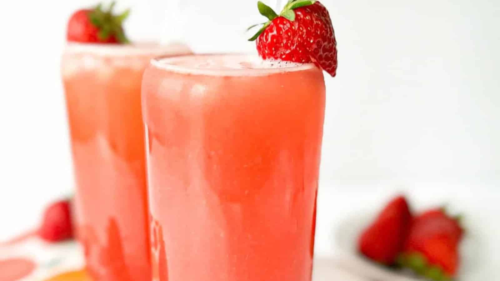 Two tall glasses filled with pink strawberry drink, each garnished with a whole strawberry on the rim; blurred strawberries in the background.