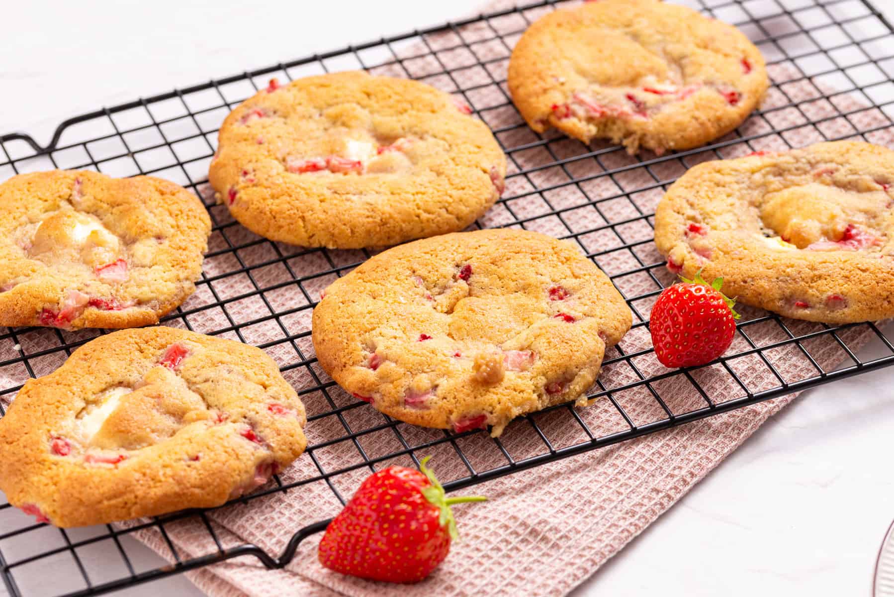 Six strawberry cookies and two fresh strawberries on a cooling rack placed over a pink textured cloth.