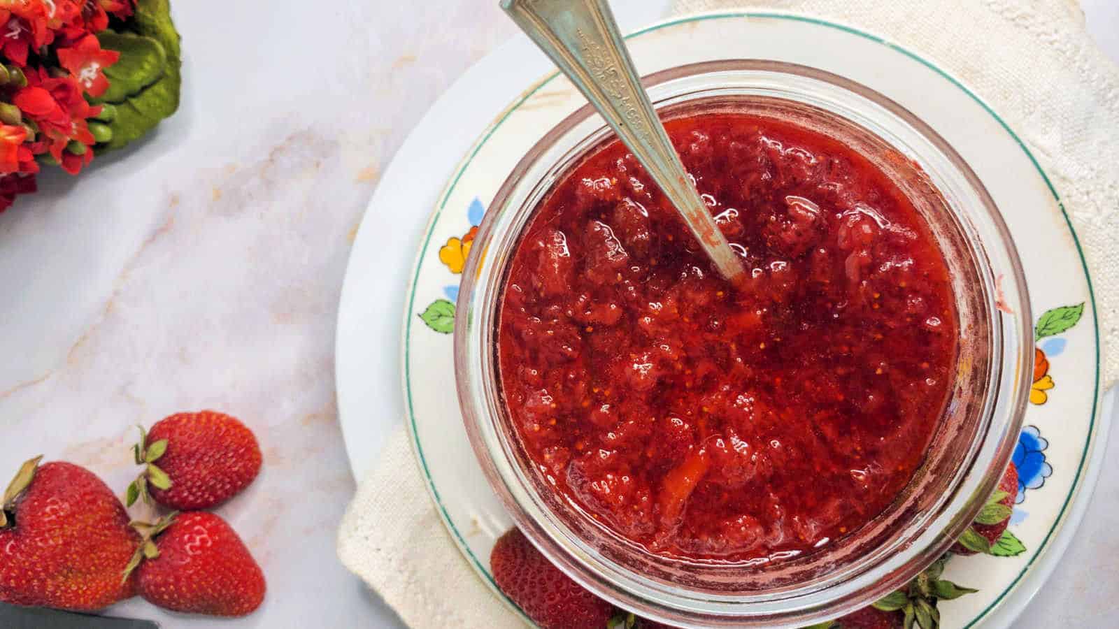 A glass jar filled with strawberry jam and a spoon, placed on a plate with a floral design, next to fresh strawberries and a cloth napkin.