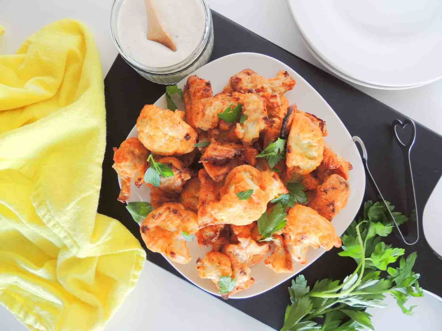 A plate of baked buffalo cauliflower bites garnished with parsley, next to a jar of dipping sauce, a yellow cloth, and fresh herbs on a black tray.