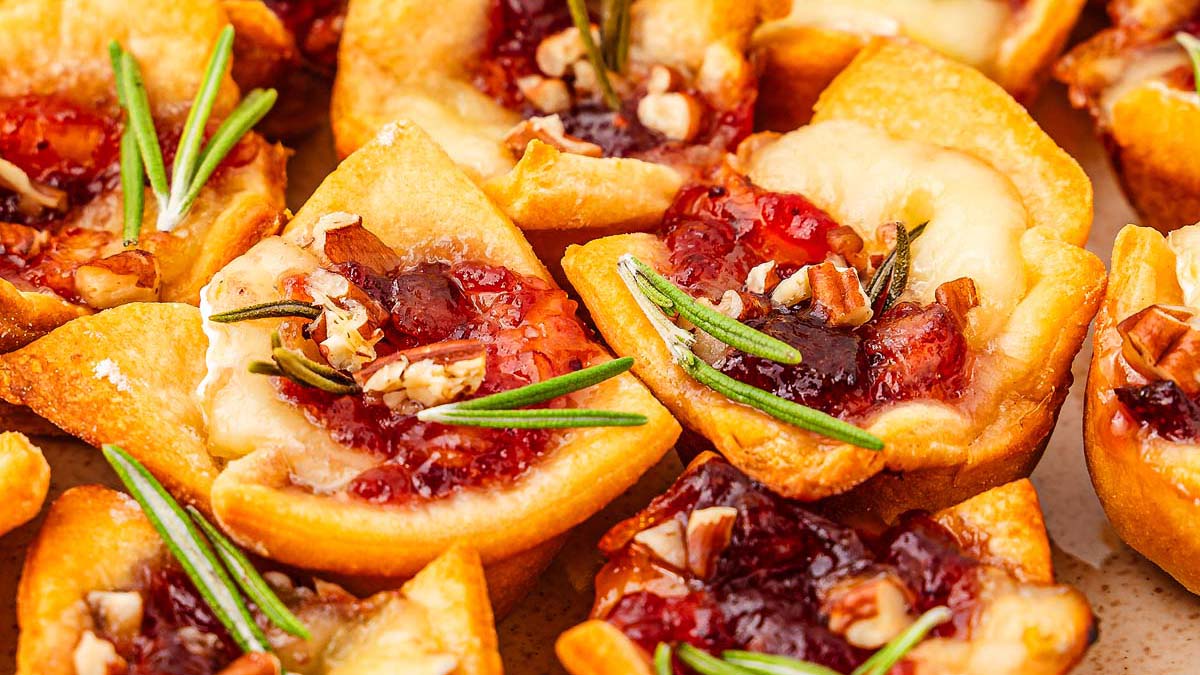 Mini baked pastry cups filled with brie cheese, cranberry sauce, chopped nuts, and garnished with sprigs of fresh rosemary.