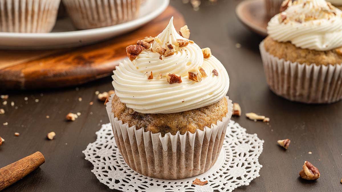 A frosted cupcake topped with chopped nuts sits on a paper doily, with more cupcakes and a cinnamon stick in the background.