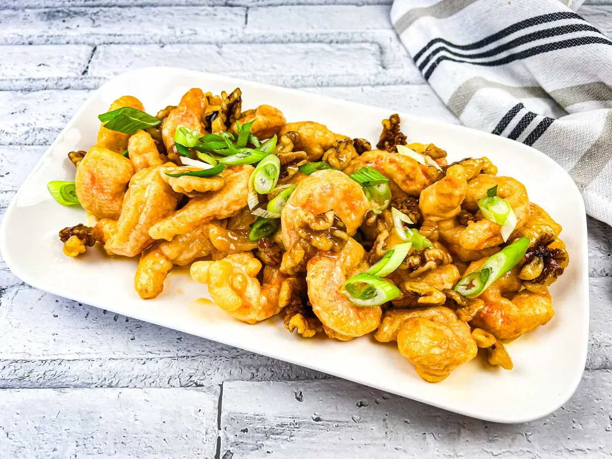 A rectangular white plate with honey walnut shrimp garnished with sliced green onions, placed on a light gray tile surface next to a striped cloth napkin.