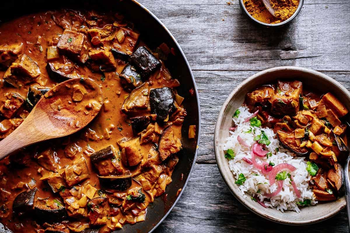 A skillet of eggplant curry with a wooden spoon next to a bowl of white rice topped with curry and herbs on a rustic wooden surface.
