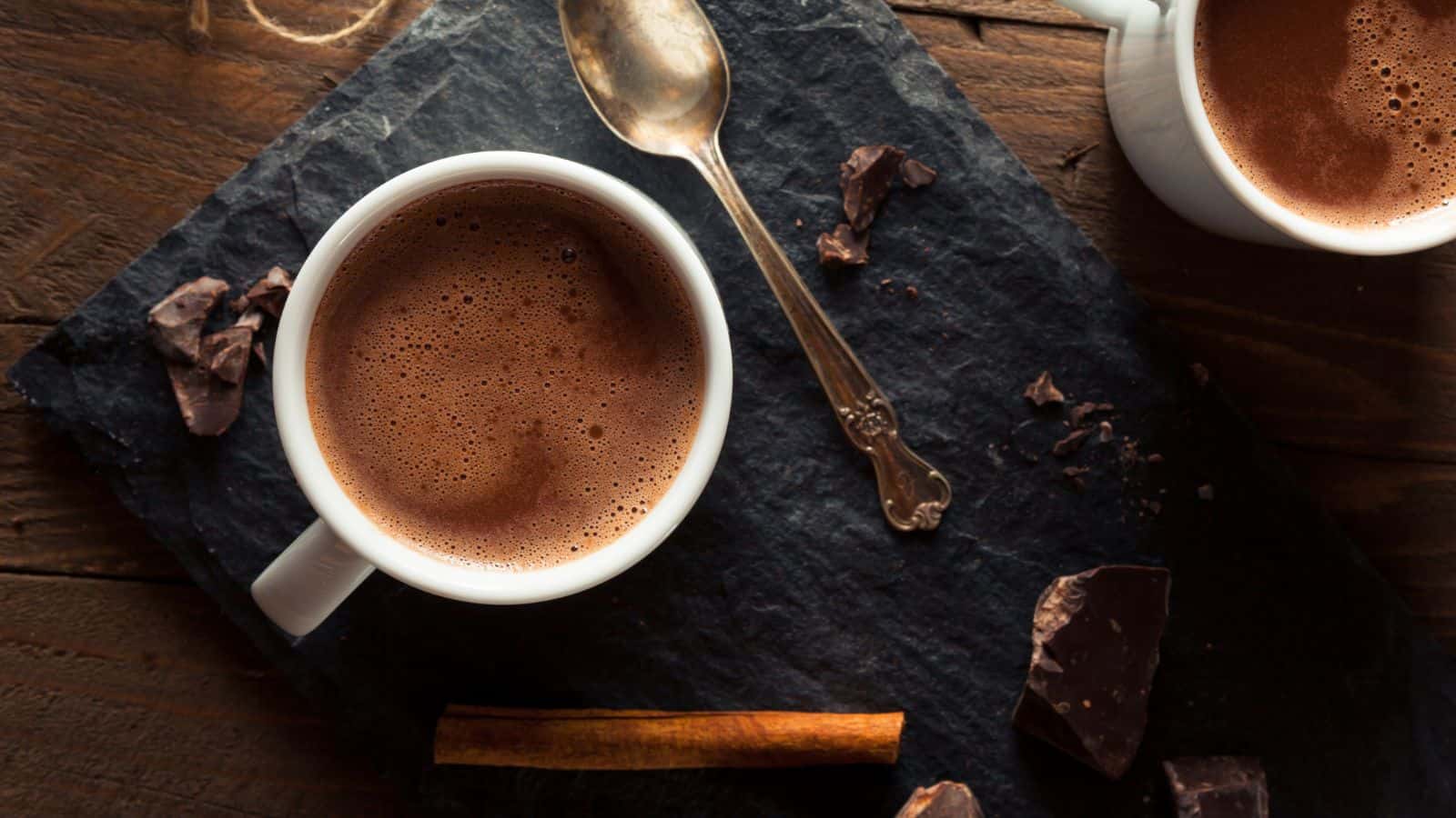A white cup filled with hot chocolate sits on a dark slate board with a silver spoon, chocolate pieces, and a cinnamon stick nearby.