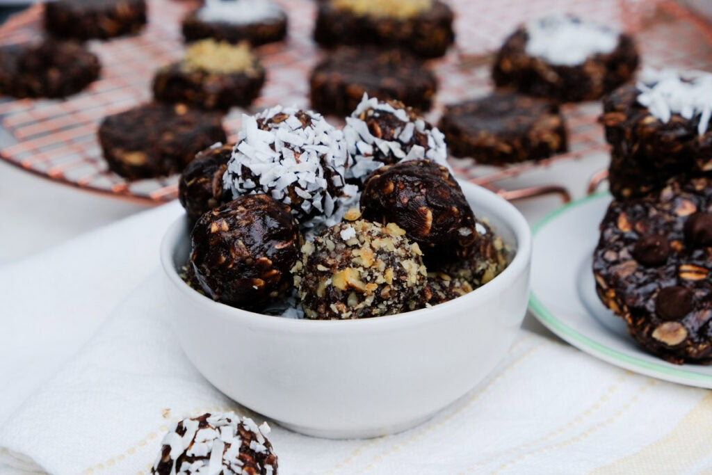 A white bowl filled with chocolate oat balls topped with coconut and chopped nuts, with more oat balls on a plate and cooling rack in the background.