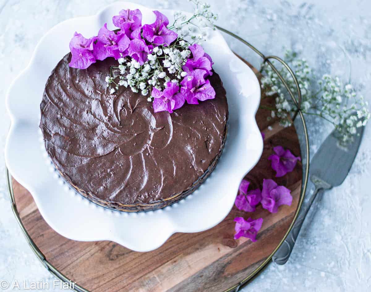 A chocolate cake topped with purple flowers and babys breath on a white cake stand, placed on a wooden board with a cake server nearby.