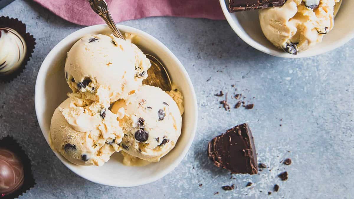 Two bowls of chocolate chip ice cream on a gray surface, one with a spoon; pieces of chocolate and truffles are nearby.