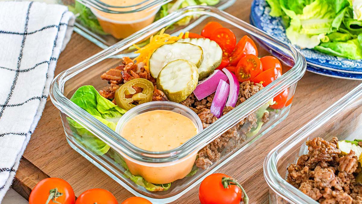 A glass container holds ground beef, lettuce, cherry tomatoes, pickles, red onion, cheese, bacon, jalapenos, and a cup of sauce, arranged as a salad.