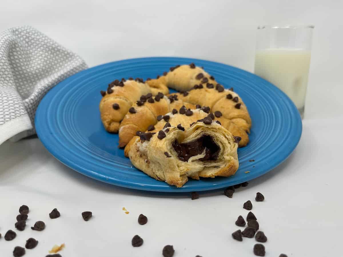 A blue plate with chocolate chip croissants, one cut open to reveal the chocolate filling, sits next to a glass of milk and a napkin. Chocolate chips are scattered on the surface.