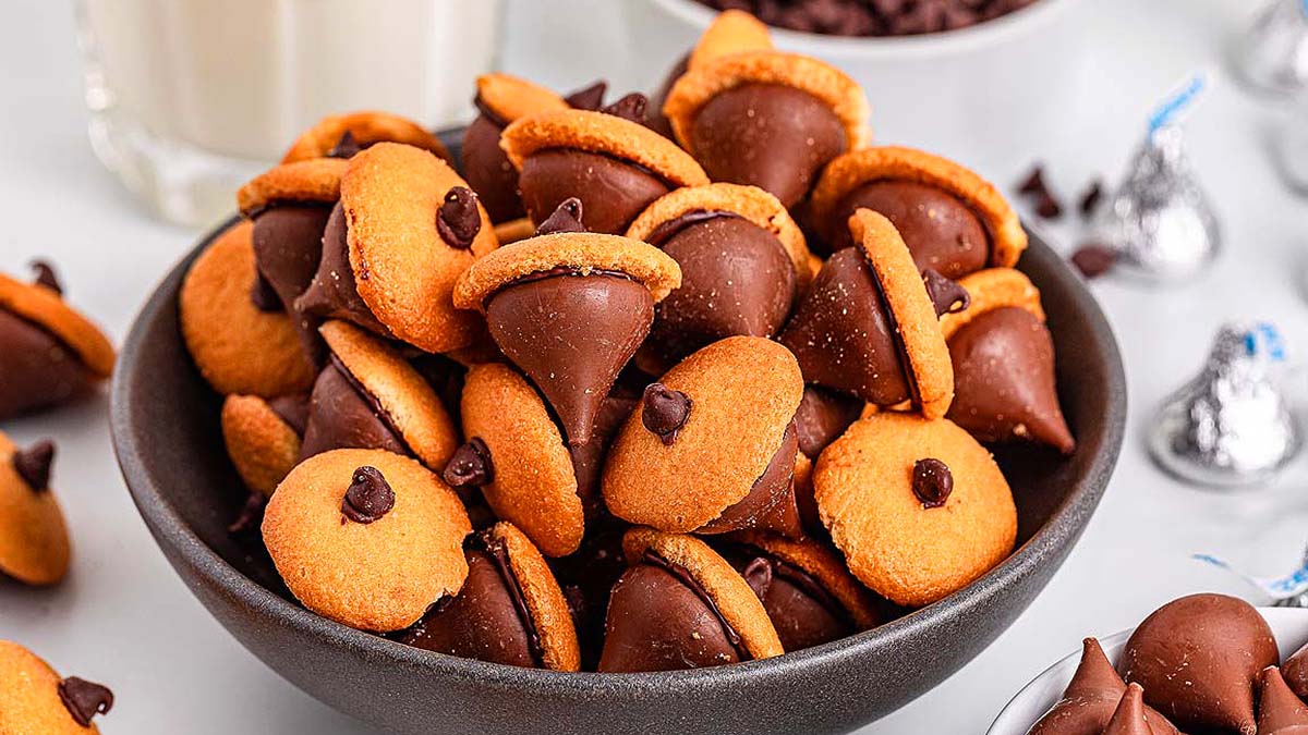 A bowl filled with acorn-shaped treats made from chocolate kisses, mini chocolate chips, and round cookies, with more treats and a glass of milk in the background.