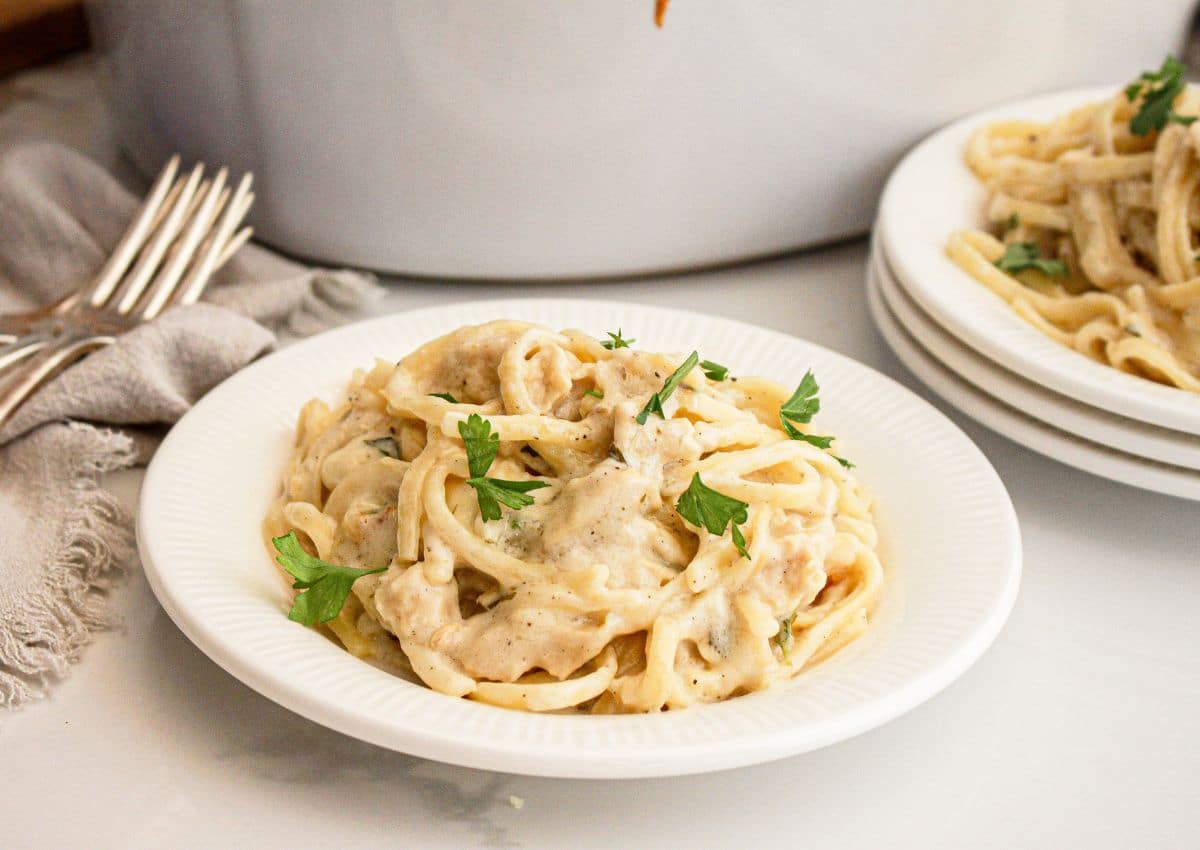 A white plate of creamy chicken alfredo pasta garnished with parsley sits on a table beside stacked plates and metal forks.