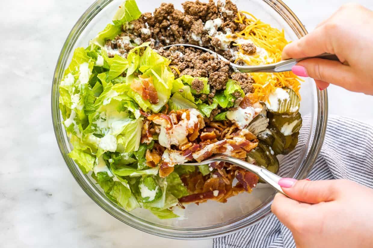 A person tossing a salad in a glass bowl with lettuce, ground beef, shredded cheese, bacon, pickles, and dressing.