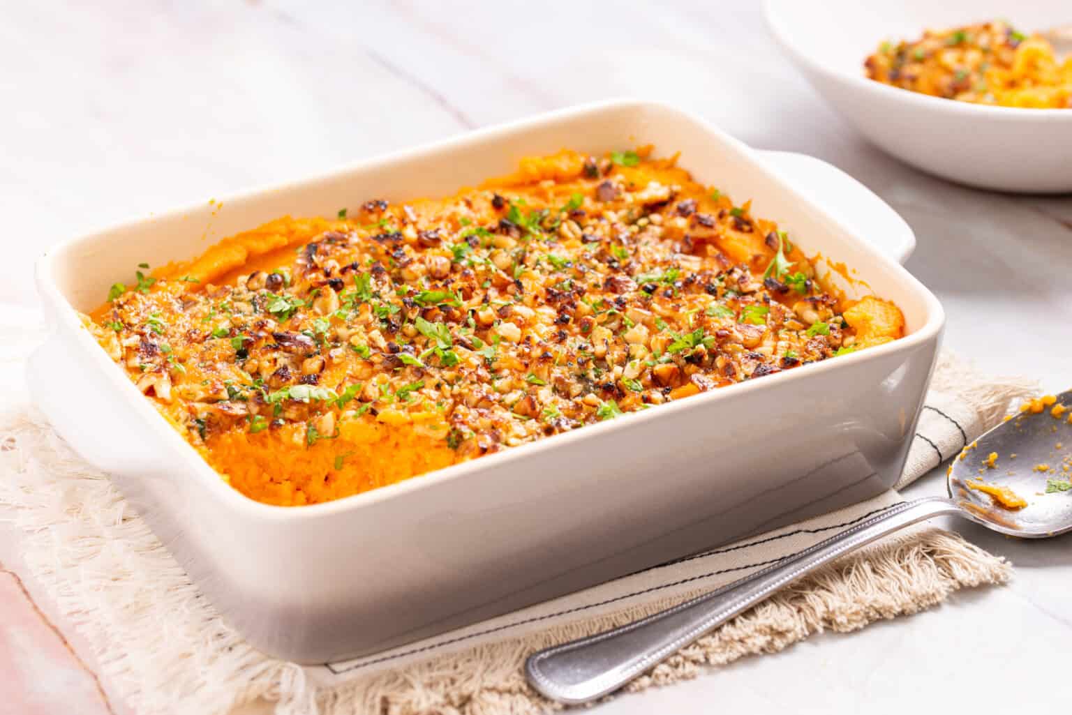 Baked casserole with a golden, textured topping and chopped herbs in a white rectangular dish, placed on a cloth napkin beside a serving spoon.