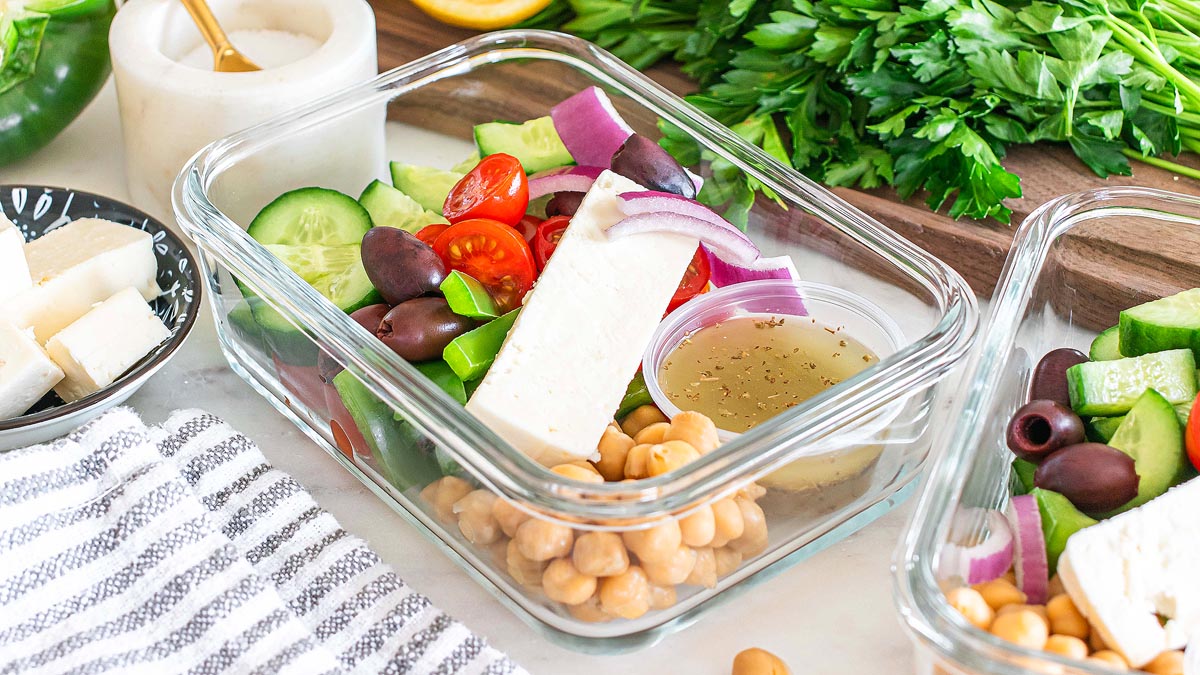 A glass container filled with Greek salad ingredients including feta, cherry tomatoes, cucumber, red onion, olives, chickpeas, and a small cup of dressing.