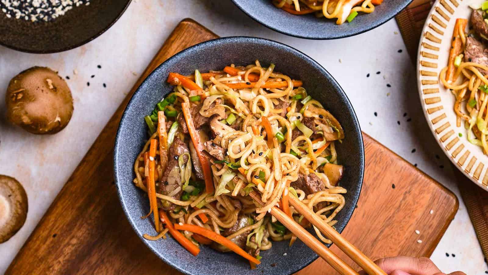 A hand uses chopsticks to pick up stir-fried noodles with beef, carrots, and vegetables from a black bowl on a wooden board.