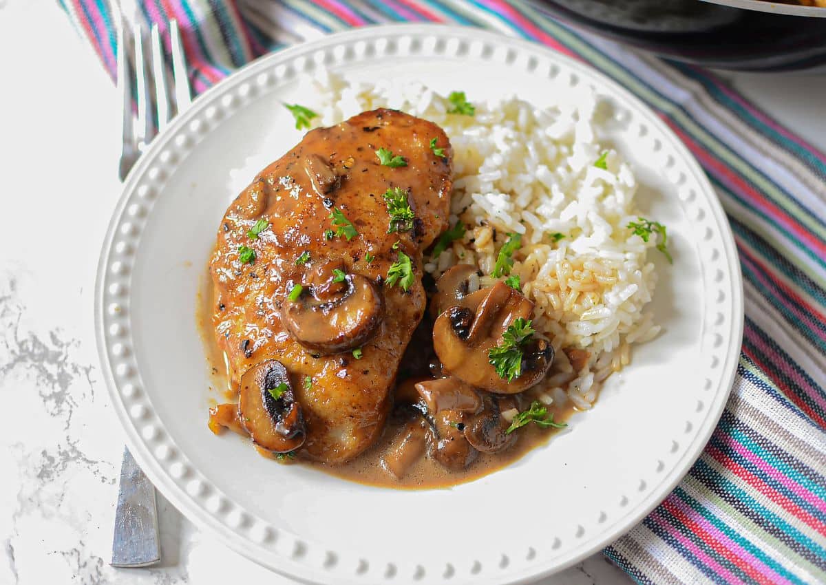 A plate with chicken breast topped with mushrooms and sauce, served alongside white rice, garnished with parsley.