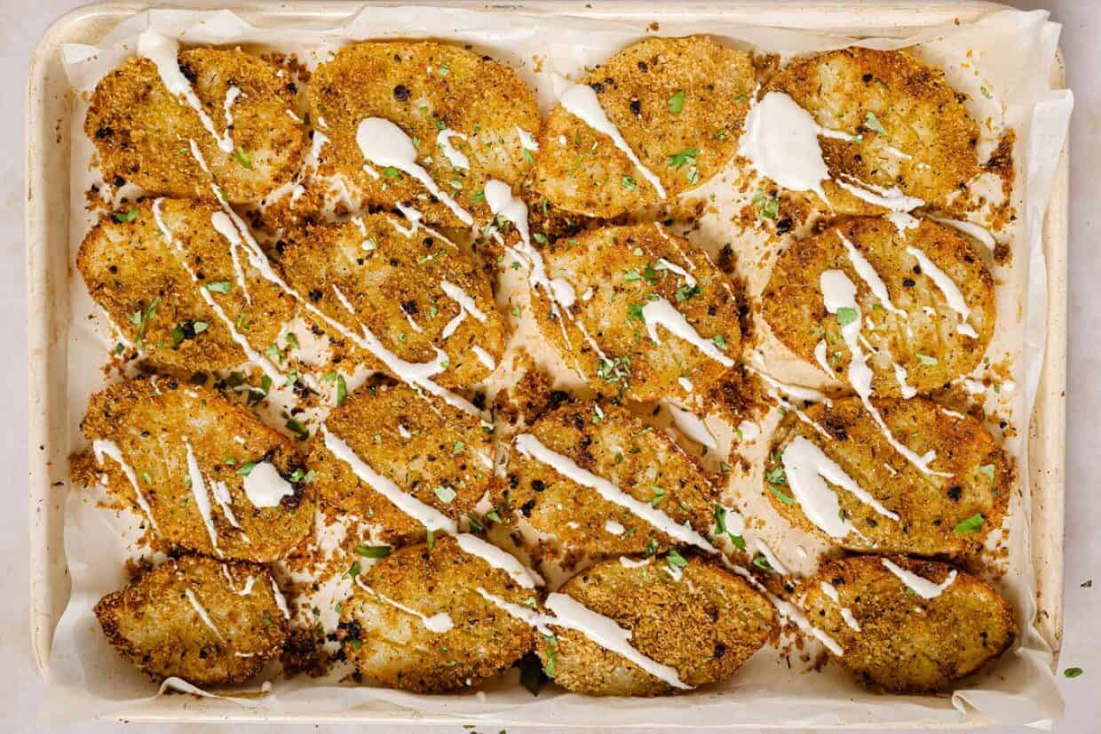 A baking tray lined with parchment holds breaded, baked slices of vegetables, lightly drizzled with a white sauce and sprinkled with chopped herbs.