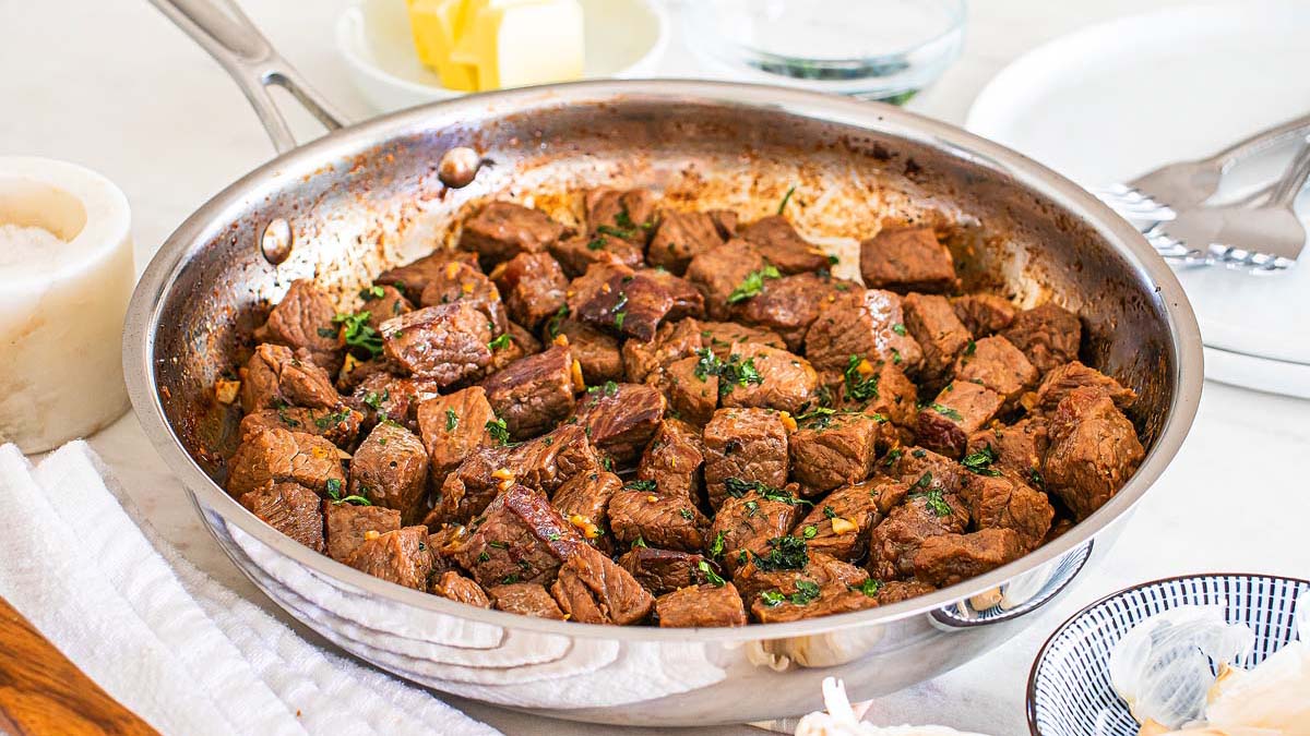 A stainless steel pan filled with cooked, seasoned beef cubes garnished with chopped herbs, placed on a kitchen counter with butter, salt, and plates nearby.