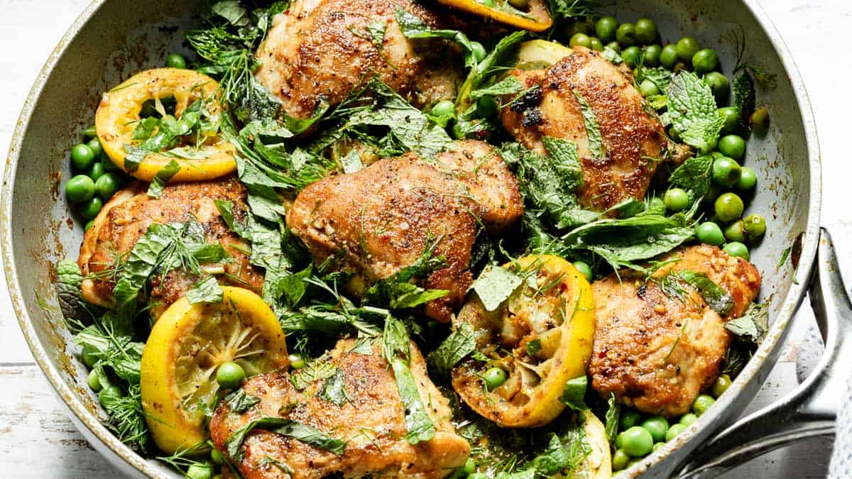 A skillet containing browned chicken thighs, lemon slices, green peas, and fresh herbs.