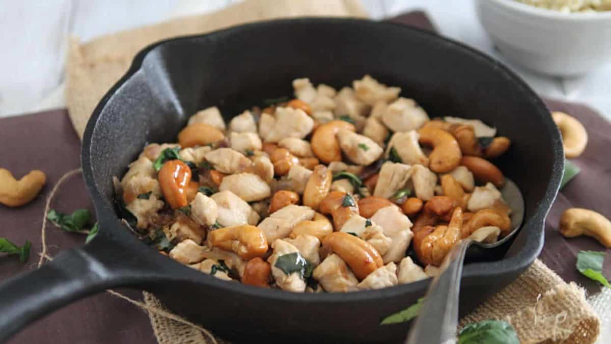 A black skillet filled with cooked diced chicken, cashew nuts, and herbs, with a spoon resting inside.