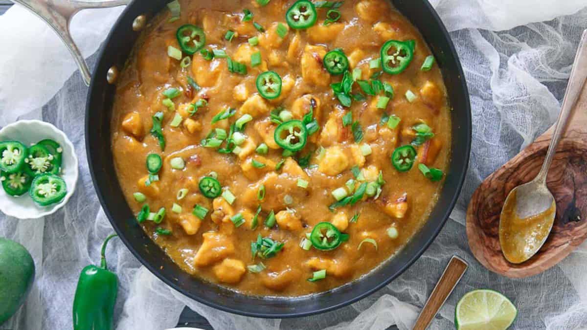 A pan filled with yellow curry containing chunks of tofu, topped with sliced green jalapeรฑos and chopped green onions, with a wooden spoon and lime wedges nearby.