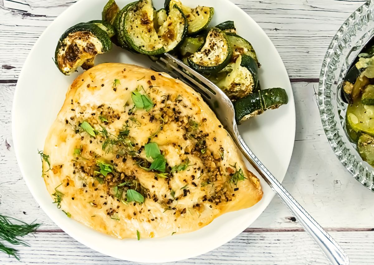 A plate with a cooked chicken breast seasoned with herbs and pepper, served with roasted zucchini slices, on a white wooden surface.