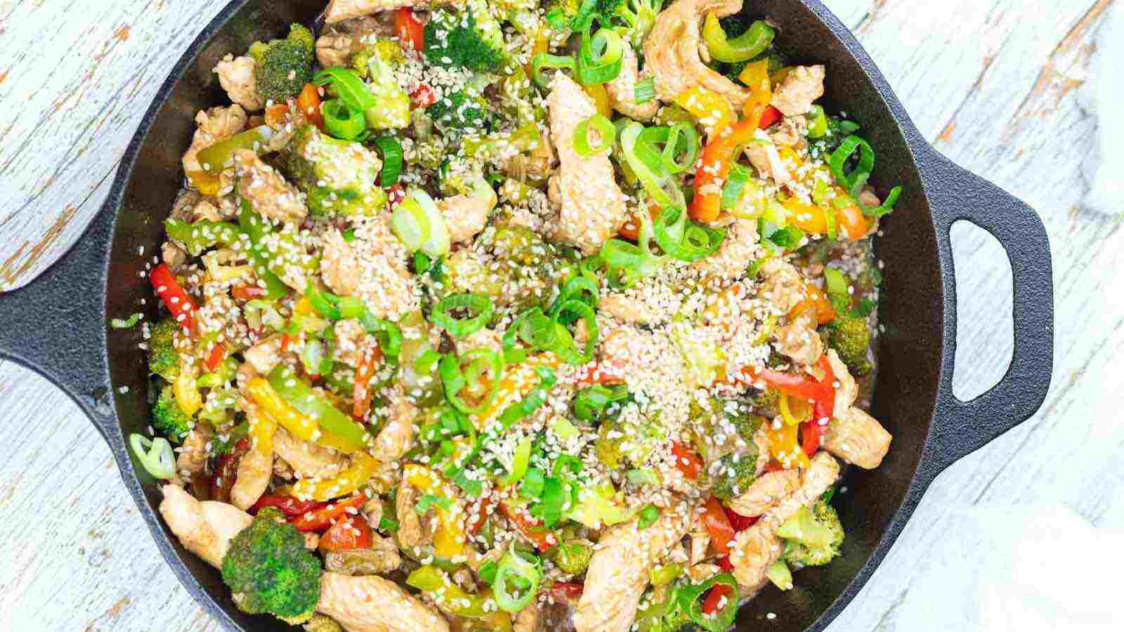A cast iron skillet filled with stir-fried chicken, broccoli, bell peppers, sliced green onions, and sprinkled with sesame seeds.