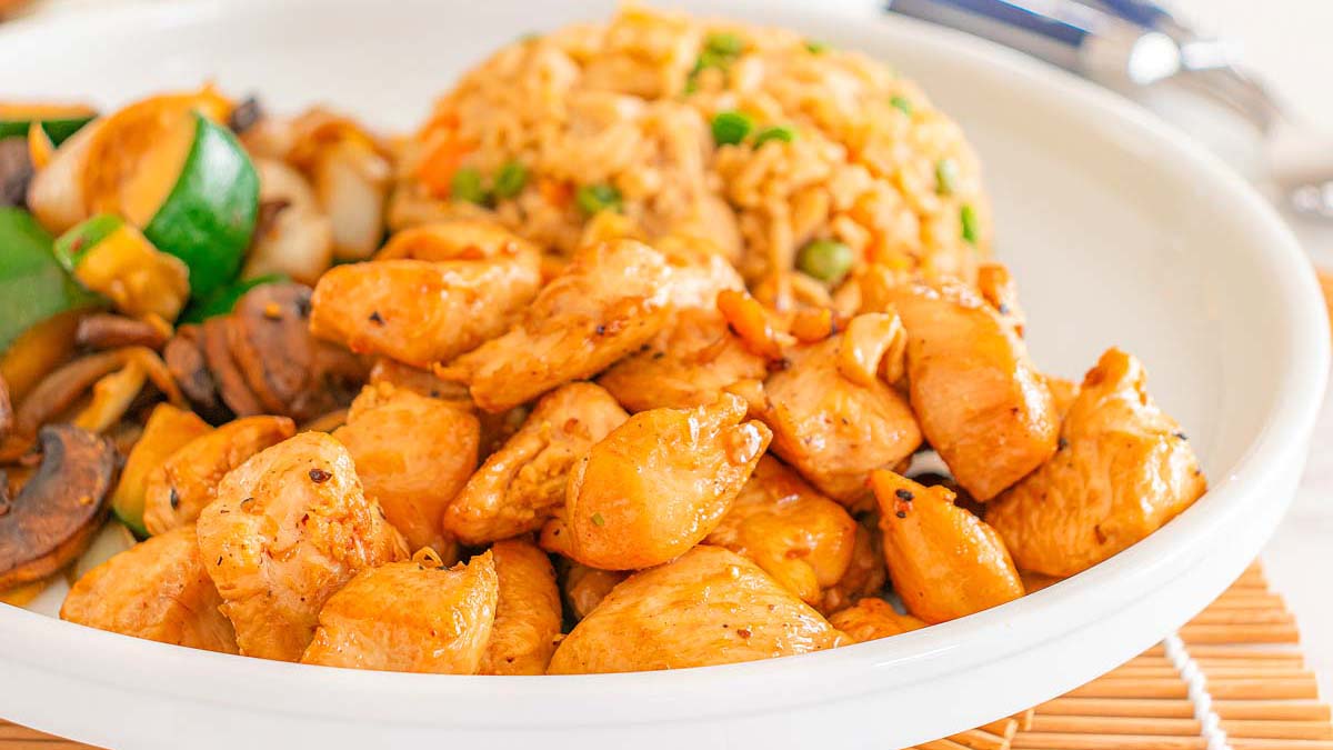 A white plate with grilled chicken pieces, stir-fried vegetables including mushrooms and zucchini, and a serving of fried rice with peas.