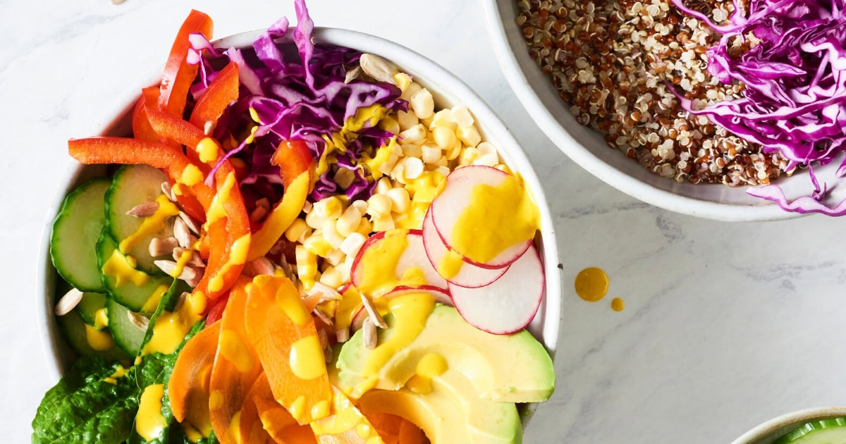 A bowl filled with sliced vegetables, avocado, corn, seeds, and a yellow dressing next to a bowl of quinoa topped with shredded purple cabbage.