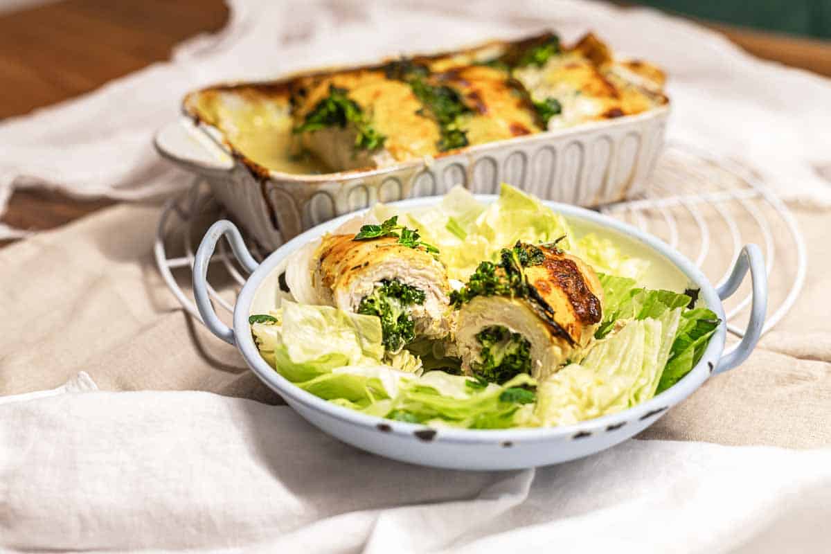 A bowl with stuffed and baked chicken breast slices on a bed of lettuce, with a baking dish of more stuffed chicken in the background.