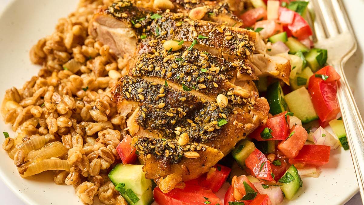 A plate with sliced seasoned chicken, a grain salad, and a mix of chopped tomatoes, cucumbers, and onions, with a fork on the side.