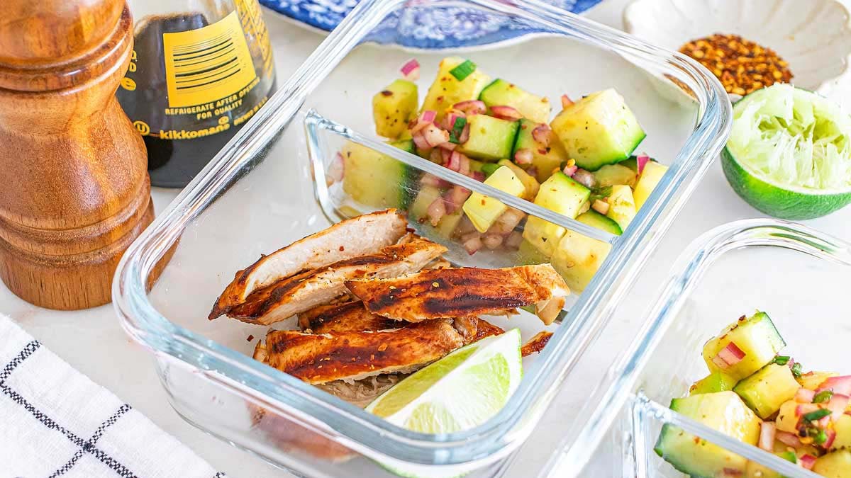 Glass meal prep container with sliced grilled chicken, a lime wedge, and cucumber salad with red onion; soy sauce, pepper grinder, and lime nearby on the table.