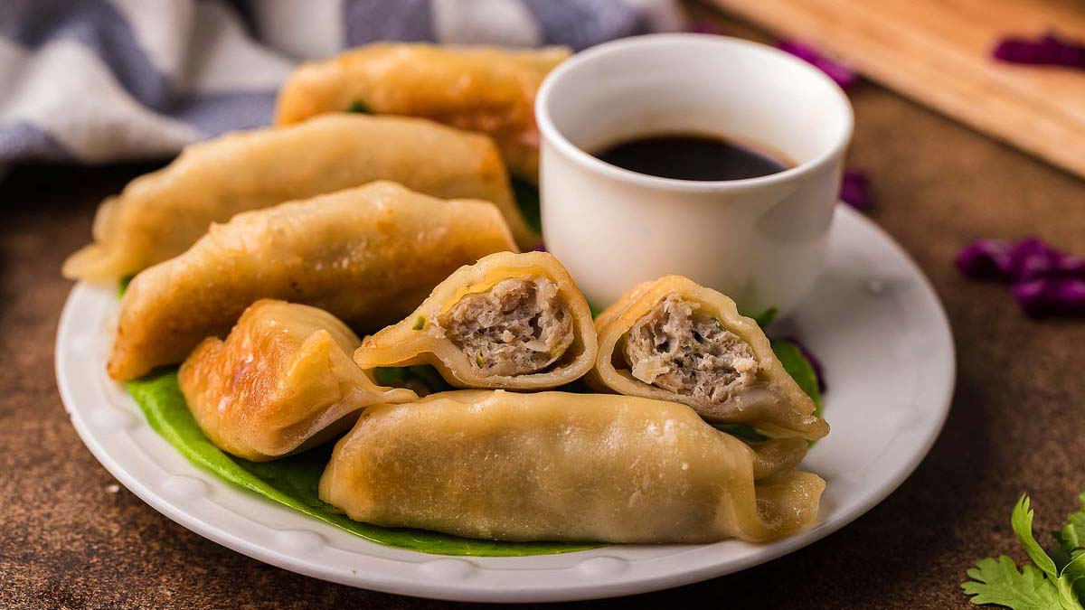 A plate of pan-fried dumplings, some cut open to show a meat filling, served with a small cup of dark dipping sauce on the side.