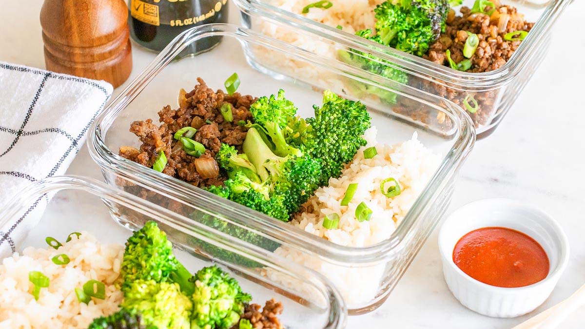 Three glass meal prep containers with rice, cooked ground beef with onions, broccoli, and chopped green onions. A small dish of red sauce and bottle of soy sauce are nearby.