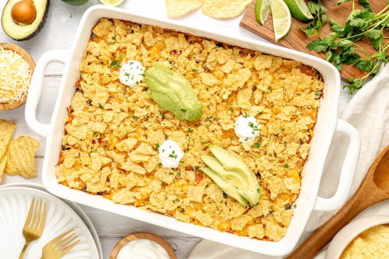 A casserole topped with crushed tortilla chips, avocado slices, and dollops of sour cream in a white baking dish, surrounded by plates, lime wedges, cilantro, and other ingredients.
