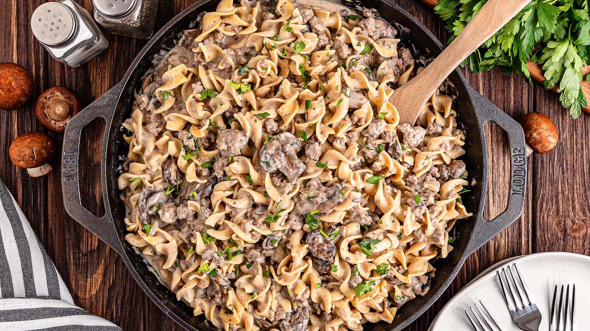 A skillet of beef stroganoff with egg noodles, mushrooms, and creamy sauce, garnished with parsley, sits on a wooden table with utensils and seasonings nearby.