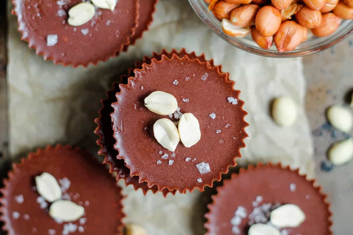 Chocolate cups topped with sea salt and peanuts are arranged on parchment paper, with a bowl of shelled peanuts nearby.
