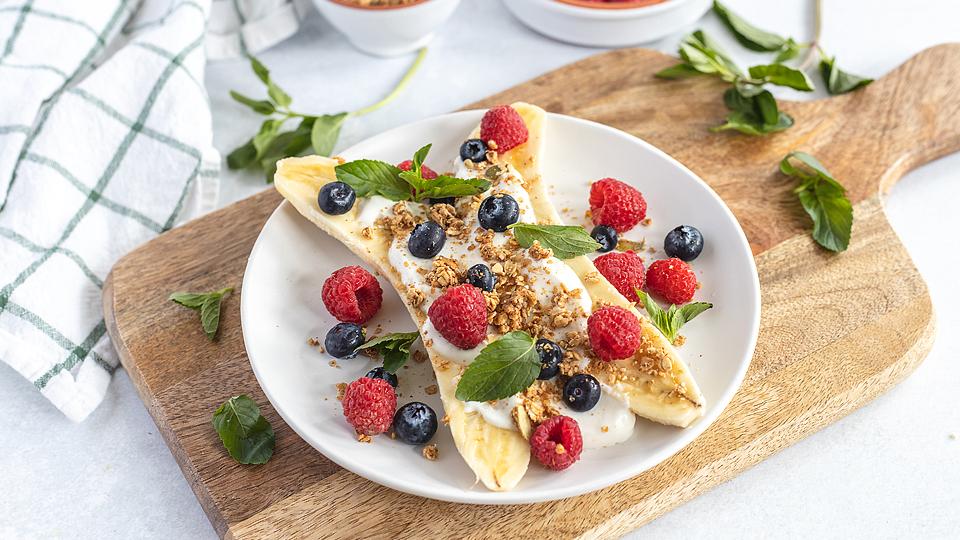 A plate with a split banana topped with yogurt, granola, raspberries, blueberries, and fresh mint, set on a wooden board.