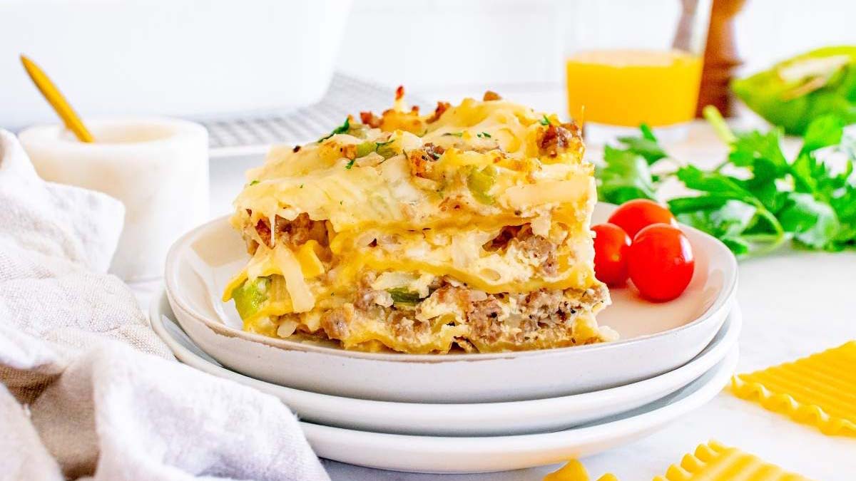 A slice of lasagna with layers of pasta, meat, and cheese is served on a white plate, garnished with herbs; tomatoes and greens are in the background.