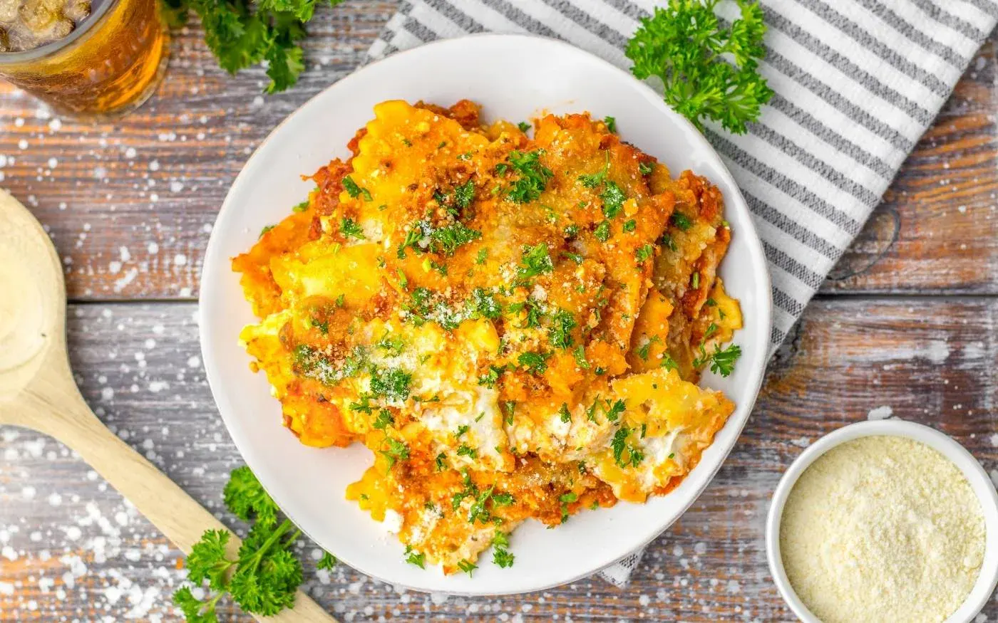 A plate of baked lasagna topped with chopped parsley and grated cheese sits on a wooden table next to a bowl of parmesan, a wooden spoon, and a striped napkin.
