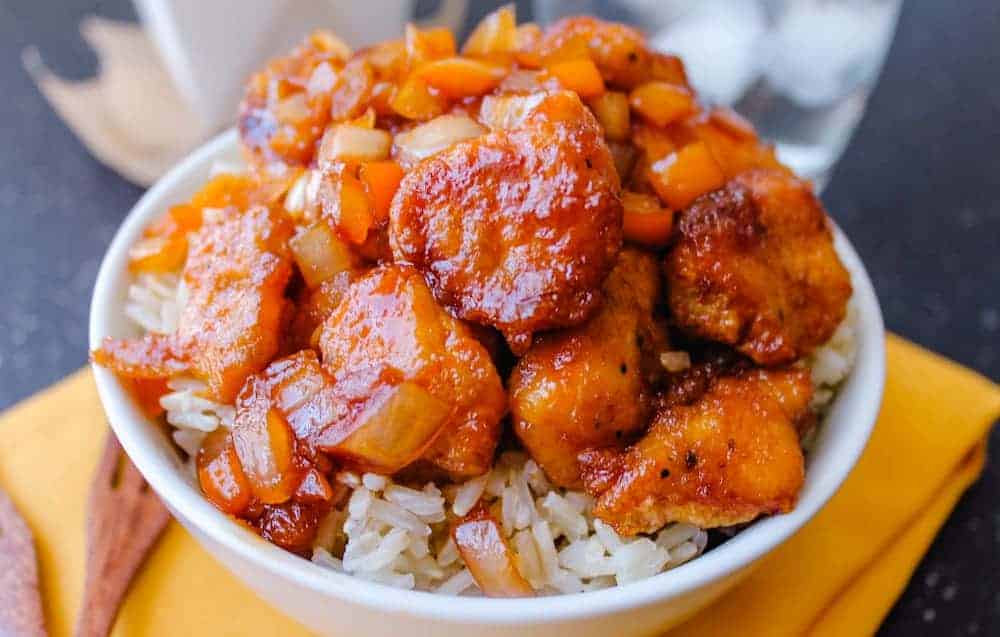 A bowl of white rice topped with glazed, fried chicken pieces, diced onions, and orange bell peppers.