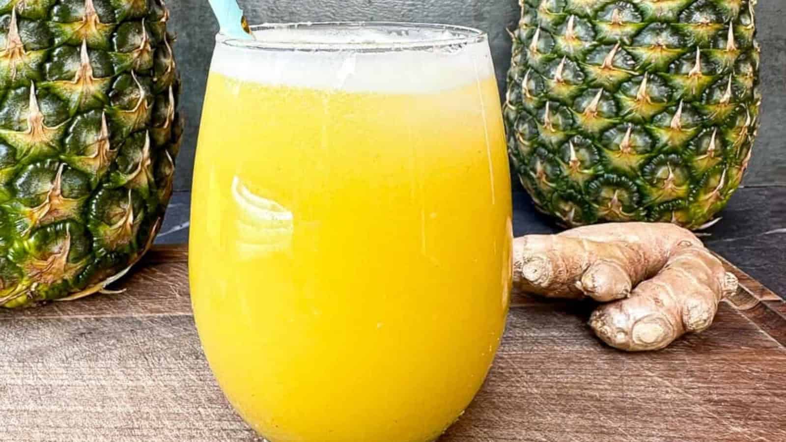 A glass of yellow juice with a straw sits on a wooden surface, flanked by two pineapples and a piece of ginger root.