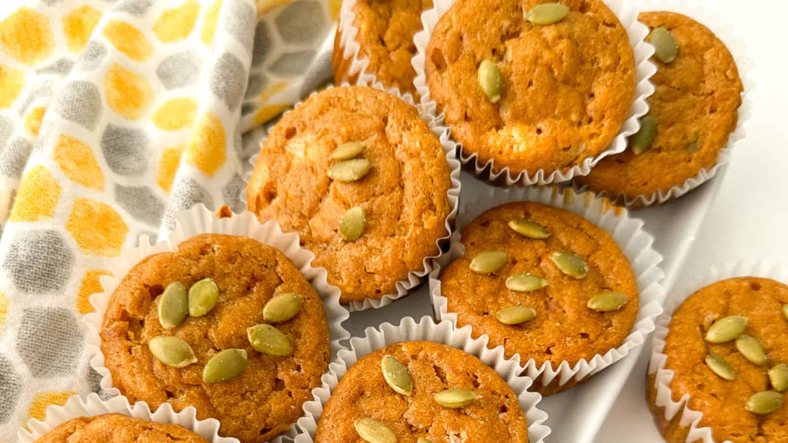 Pumpkin muffins in white paper liners, some topped with pumpkin seeds, arranged on a white plate next to a patterned yellow and gray cloth.